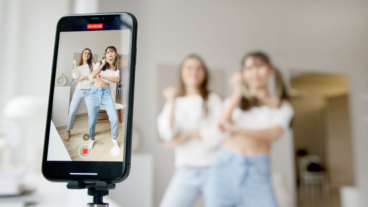 services-02 Two women are dancing indoors while filming content with a smartphone camera.